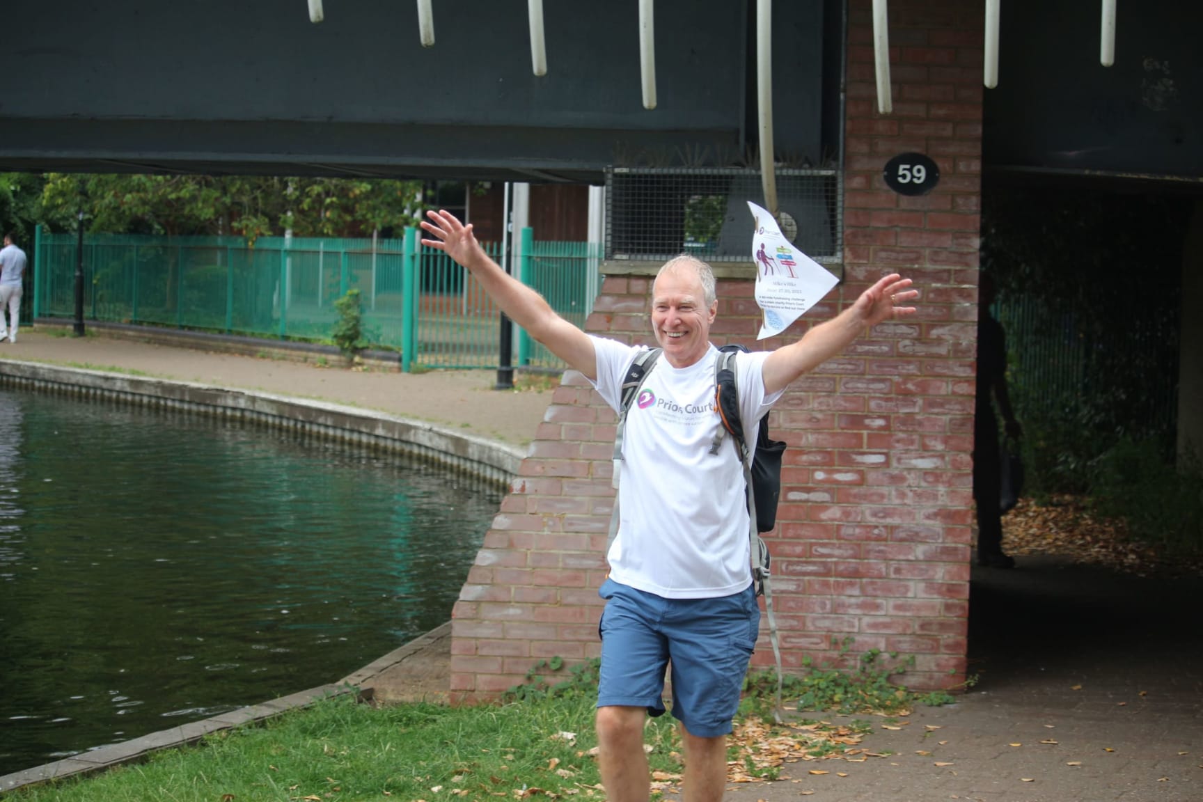 a man dressed in a white t-shirt walks under a bridge next to a canal. his arms are raised in celebration